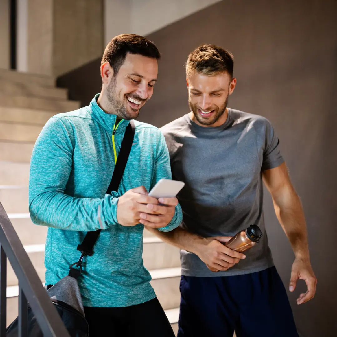 Two men in athletic wear stand on stairs, smiling at a phone. One holds a water bottle and the other carries a gym bag.